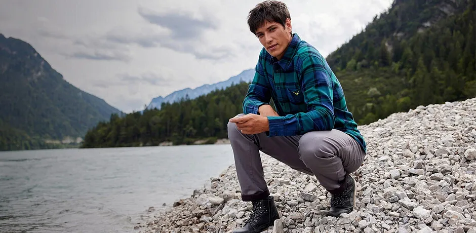 Muž v kostkované košili a šedých kalhotách sedí na břehu řeky v horské krajině.
