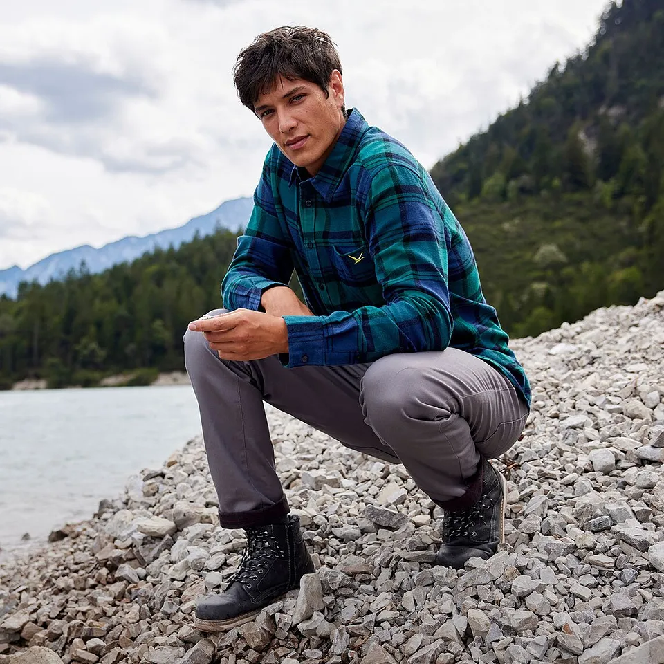 Muž v kostkované košili a šedých kalhotách sedí na břehu řeky v horské krajině.