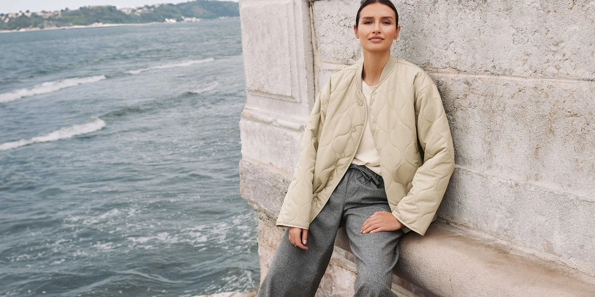 Ležérní podzimní look: béžová bunda, světle modré džíny a bílé tričko u vody.