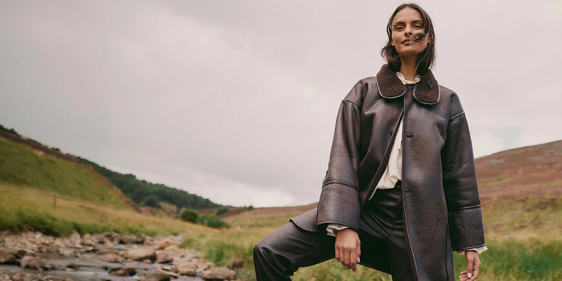 Elegantní hnědý kožený kabát s velkým límcem v kombinaci se světlým topem a tmavými kalhotami.