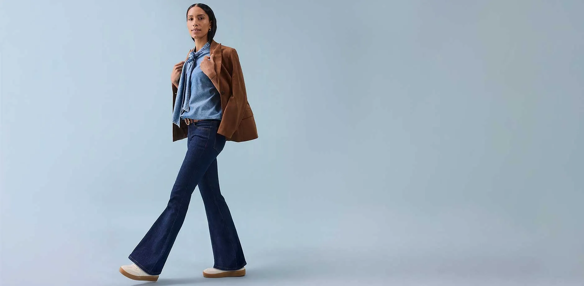 Žena má na sobě zvonové kalhoty, top bez rukávů a hnědou bundu nonšalantně přehozenou přes rameno.