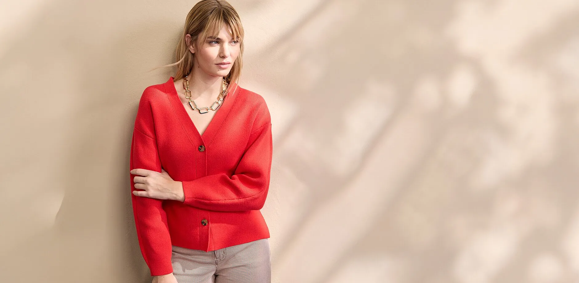 Z&aacute;řivě červen&yacute; svetr s v&yacute;střihem do V a knofl&iacute;ky v kombinaci s n&aacute;padn&yacute;m zlat&yacute;m řet&iacute;zkem.