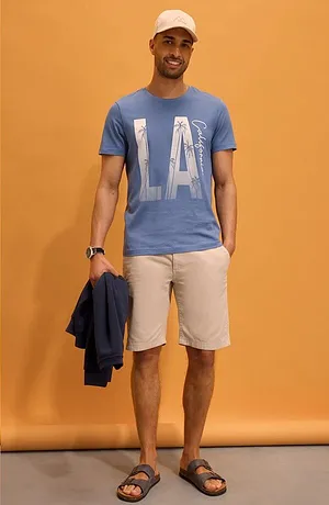 Modré tričko s nápisem „LA“, světlé šortky, bílá čepice a sandály. Ležérní letní look.