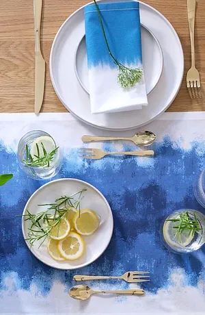 Stůl zdobí modrý ubrus s barevným přechodem, doplněný bílým nádobím a elegantními příbory.