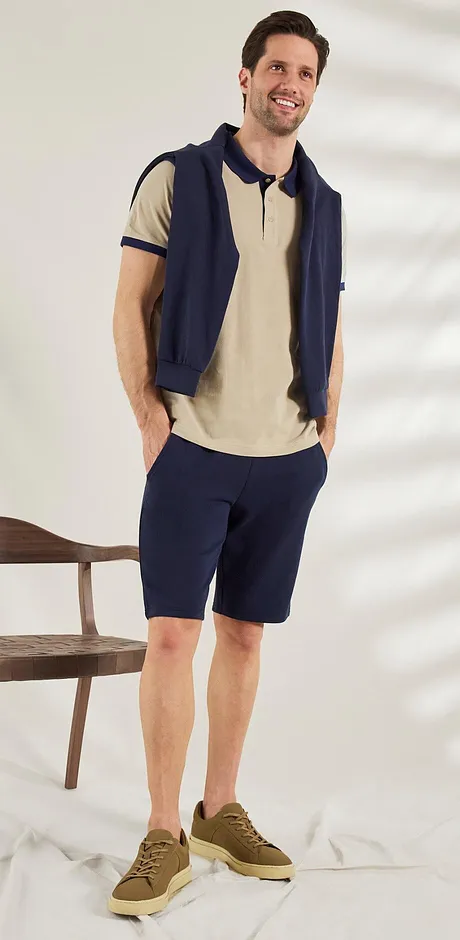 Lässiger Sommerlook: Beige Poloshirt, dunkelblaue Weste und Shorts, braune Sneaker. Entspannter Stil.