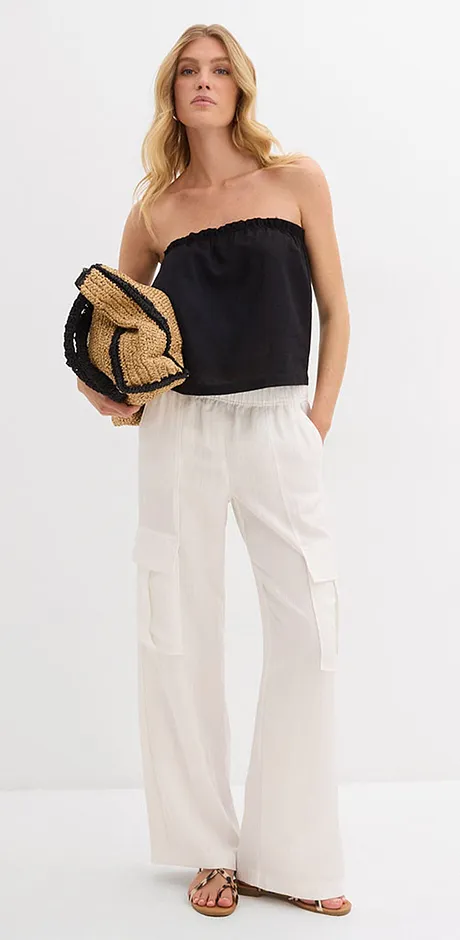 Letní outfit s černým topem bez ramínek, bílými lněnými kalhotami a pleteným slaměným kloboukem.