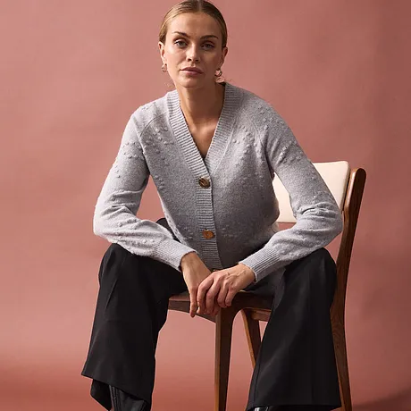Elegantní šedý svetr s velkým knoflíkem v kombinaci s černými kalhotami.