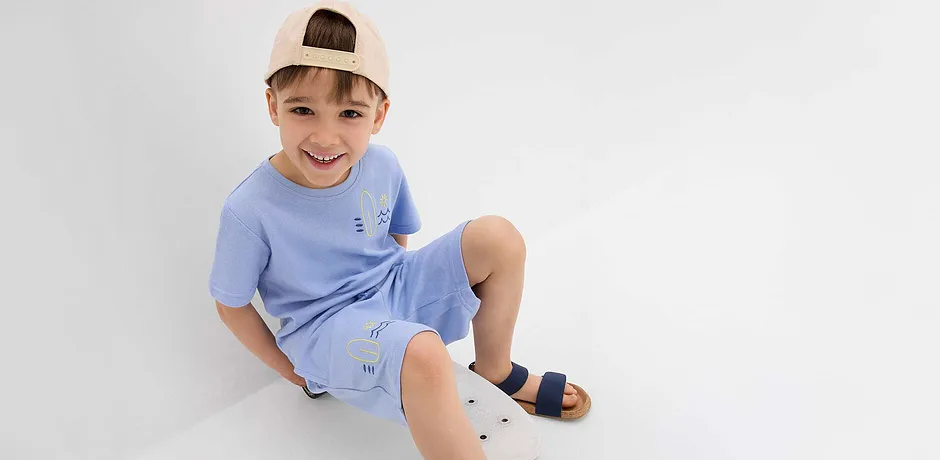 Hellblaues T-Shirt und kurze Hose mit niedlichen Stickereien, beige Baseballkappe und dunkelblaue Sandalen.