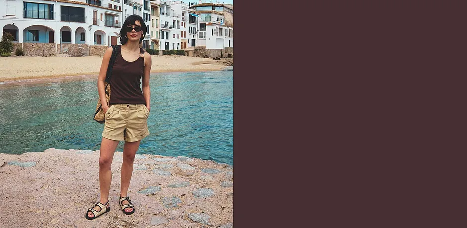 Žena v černém tílku, béžových šortkách a sandálech na pláži.