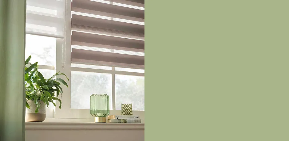 Decentní design oken s plisé v zemitých tónech. Zelená výzdoba a pokojové rostliny dodávají místnosti přirozenou atmosféru.