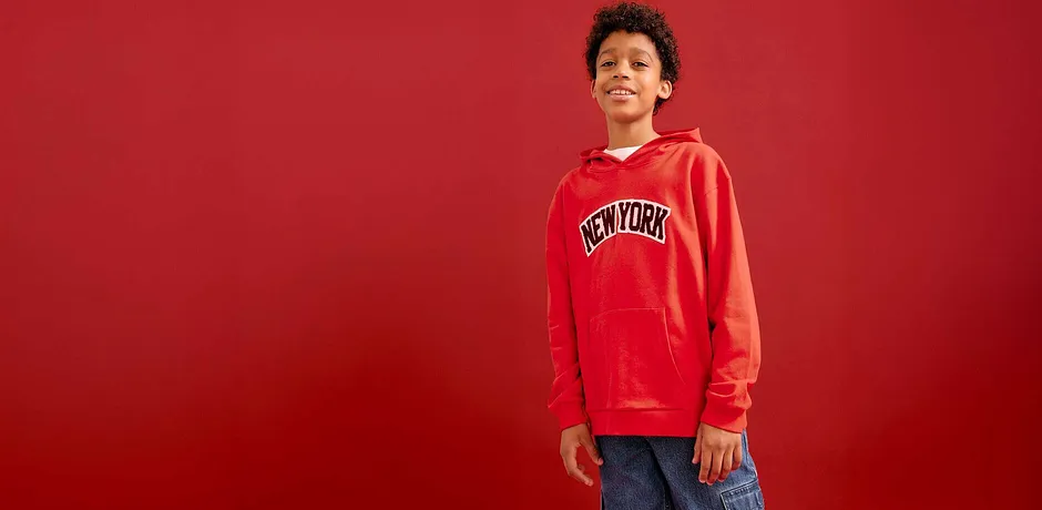 Červená mikina s kapucí a potiskem, modré džíny. Ležérní streetwearový look na červeném pozadí.