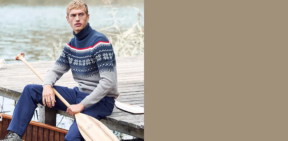 Muž v vzorovaném norském svetru a džínách sedí uvolněně na lodi.