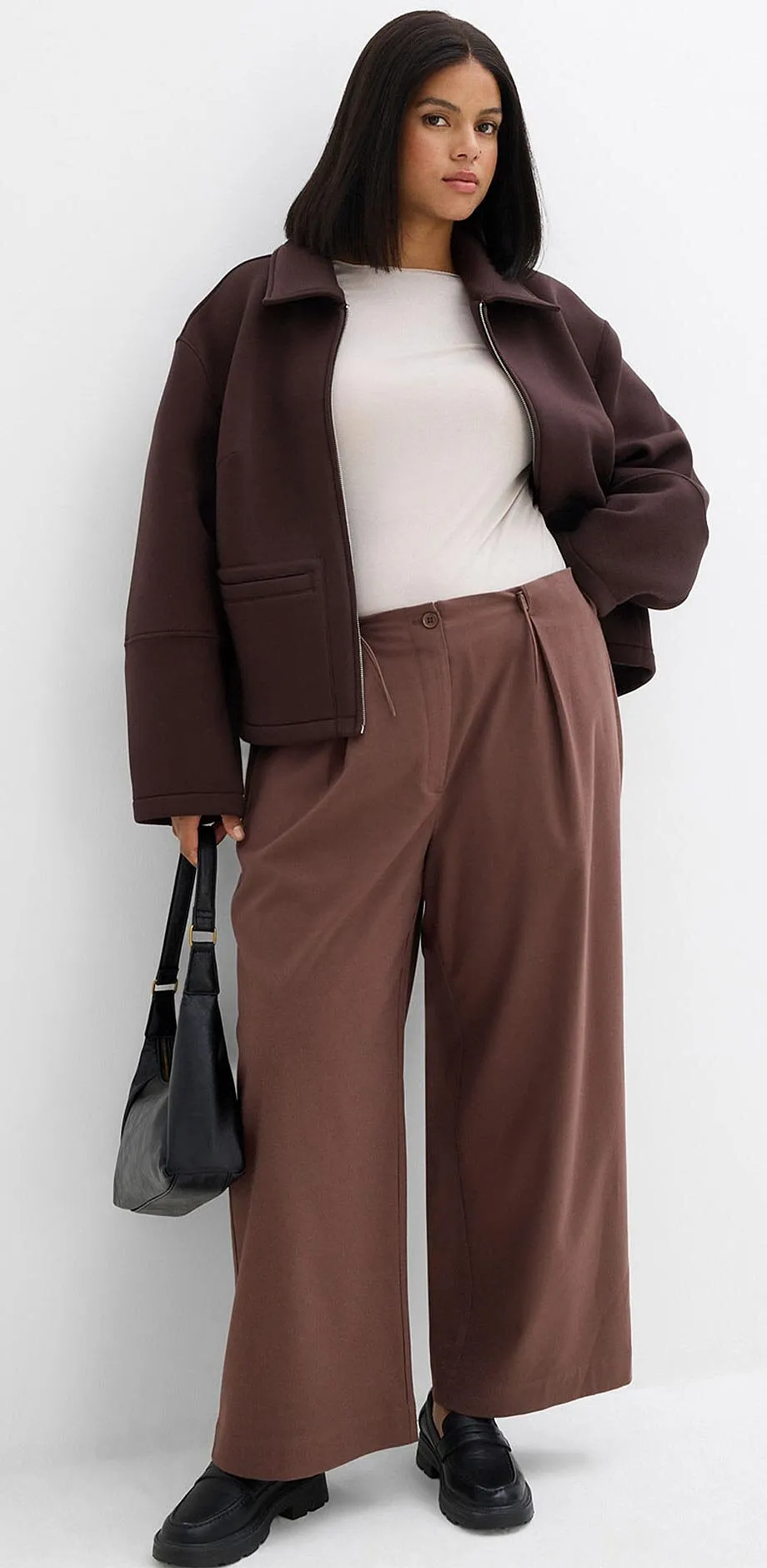 Hnědá bunda, bílé tričko a široké kalhoty v zemitých barvách.